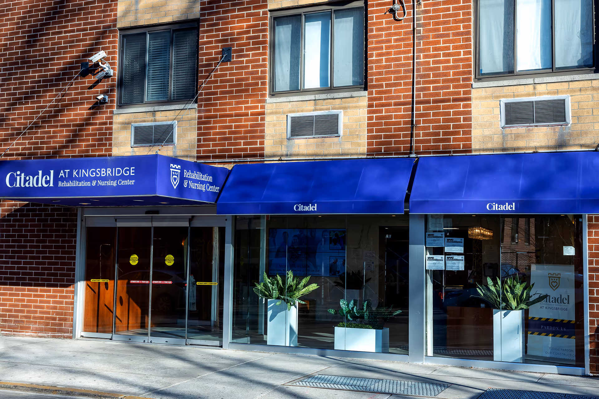 Exterior front entrance of Citadel Rehabilitation and Nursing Center at Kingsbridge with blue awnings, glass doors, and potted plants on the sidewalk in front of a brick building.