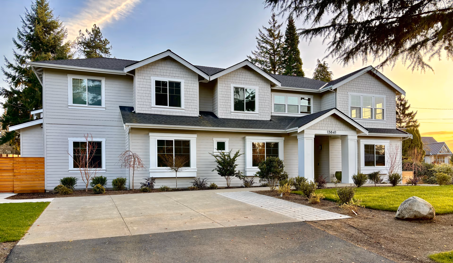Front exterior of a two-story gray suburban house with a driveway, landscaped yard, and trees at sunset.