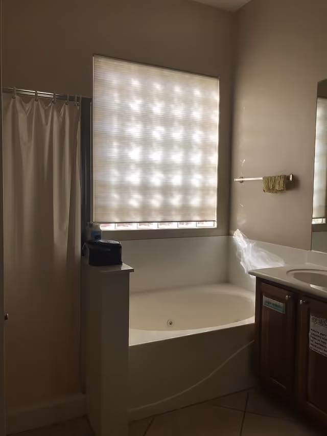 A bathroom with a large bathtub under a window covered by a translucent blind. To the left of the bathtub is a shower with a white curtain. To the right is a wooden vanity with a sink and a mirror above it. A small towel hangs on a rack on the wall above the bathtub.