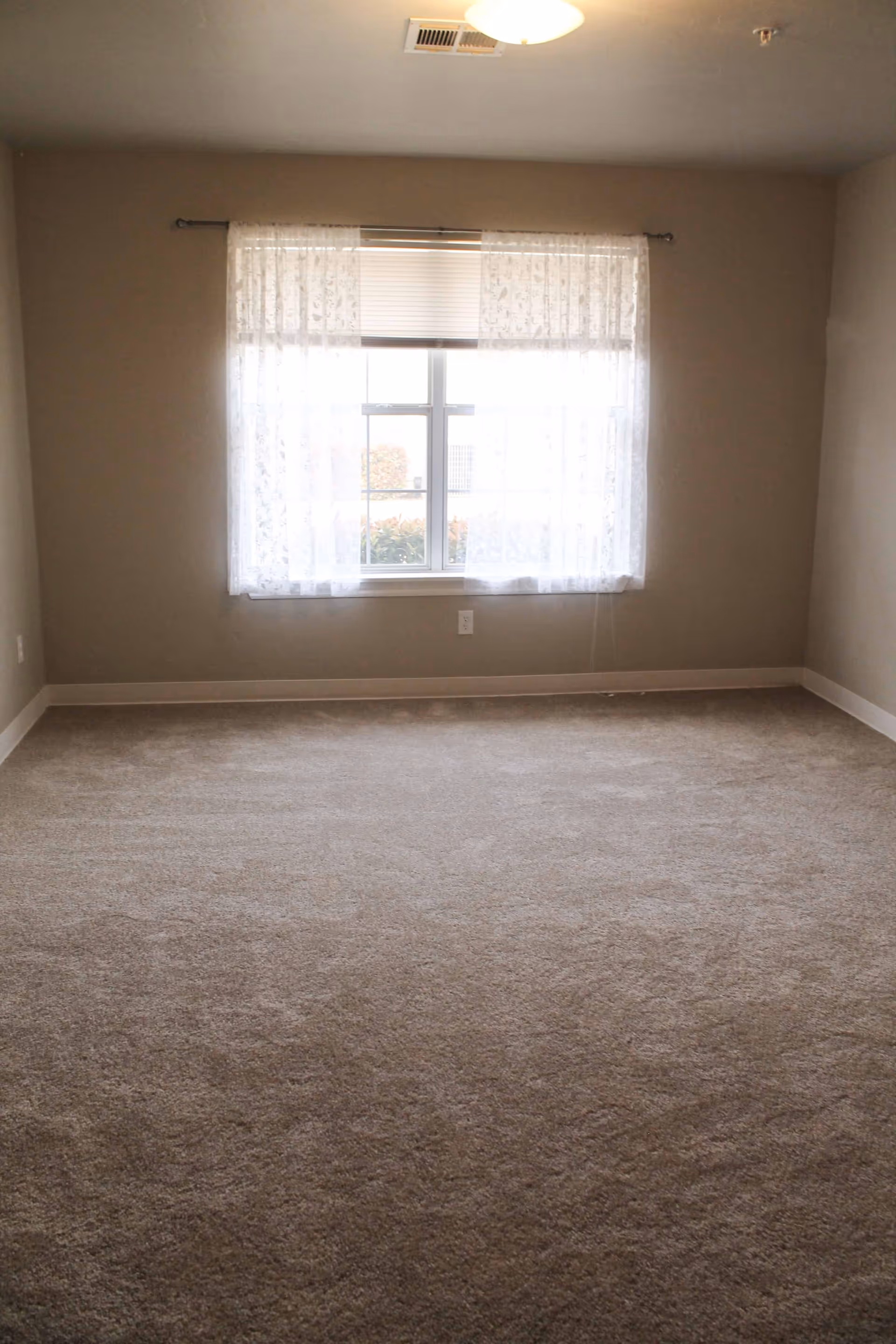 Empty room with beige carpet, beige walls, a window with sheer white curtains and a white ceiling light fixture.