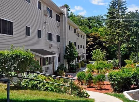 Exterior view of Springvale Apartments showing a multi-story beige building with several windows. In front of the building is a landscaped garden with bushes, trees, and a paved walkway leading through the greenery. The sky is blue with some clouds.