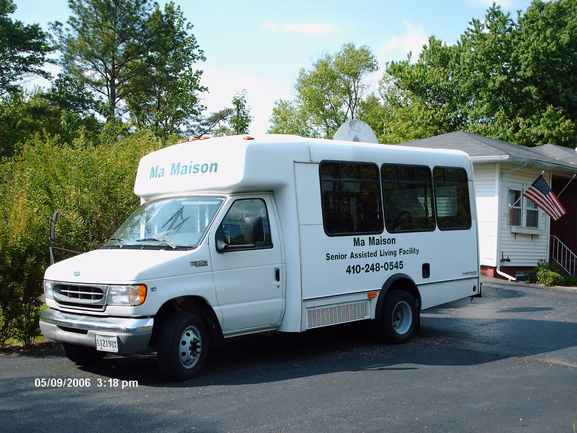 White shuttle bus parked on a driveway next to a house with an American flag. The bus has 'Ma Maison Senior Assisted Living Facility 410-248-0545' written on the side and 'Ma Maison' on the front top. Trees and bushes are visible in the background under a clear sky.