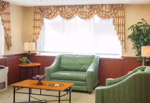 Cozy senior living lounge with green sofas, coffee table, lamps, and potted plants.