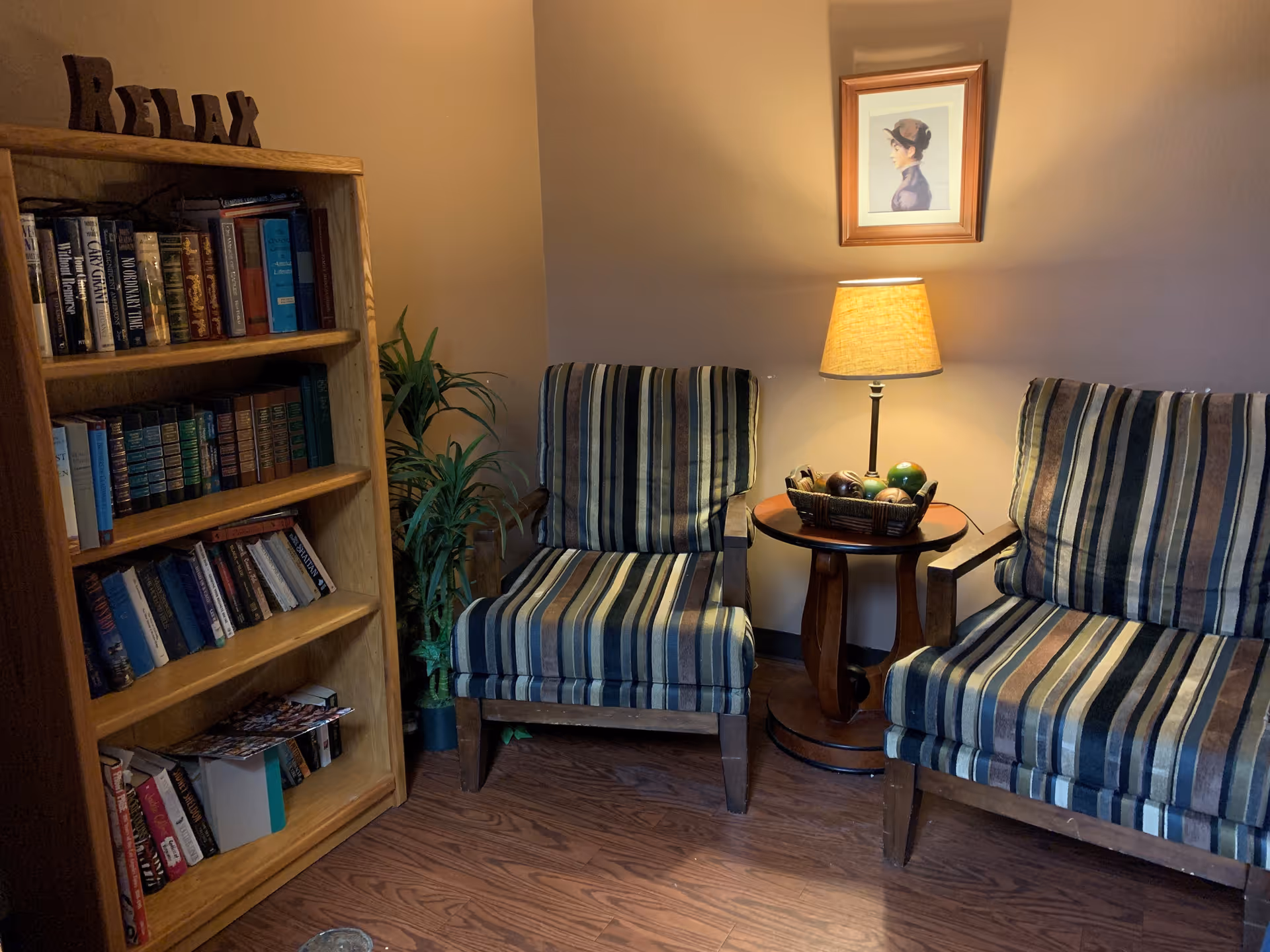A cozy corner with two striped armchairs separated by a small wooden side table holding a lamp and a basket of decorative items. To the left, there is a wooden bookshelf filled with books and a decorative 'RELAX' sign on top. A framed portrait hangs on the wall above the lamp, and a potted plant is placed between the bookshelf and the chairs. The floor is wooden.