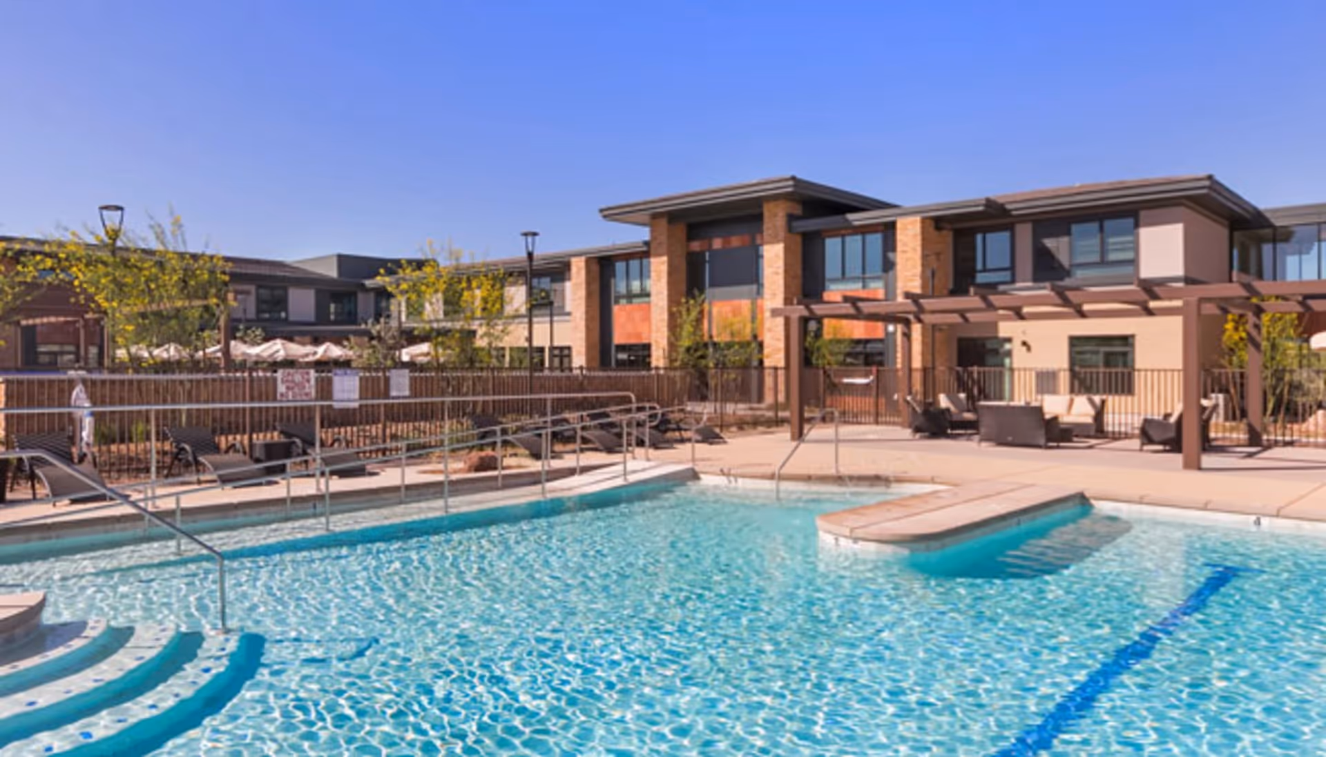 Outdoor swimming pool with clear blue water, surrounded by lounge chairs and a pergola with seating. In the background, there is a two-story building with large windows and brick accents under a clear blue sky.
