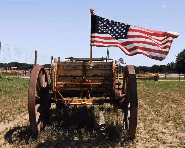 An old wooden wagon with large wooden wheels standing on a grassy field. An American flag is mounted on the wagon, waving in the wind. In the background, there is a fenced area and trees under a clear blue sky.