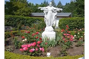 White statue of Jesus with outstretched arms standing in the center of a garden bed filled with blooming pink and red flowers, surrounded by green bushes and trees with a building roof visible in the background.