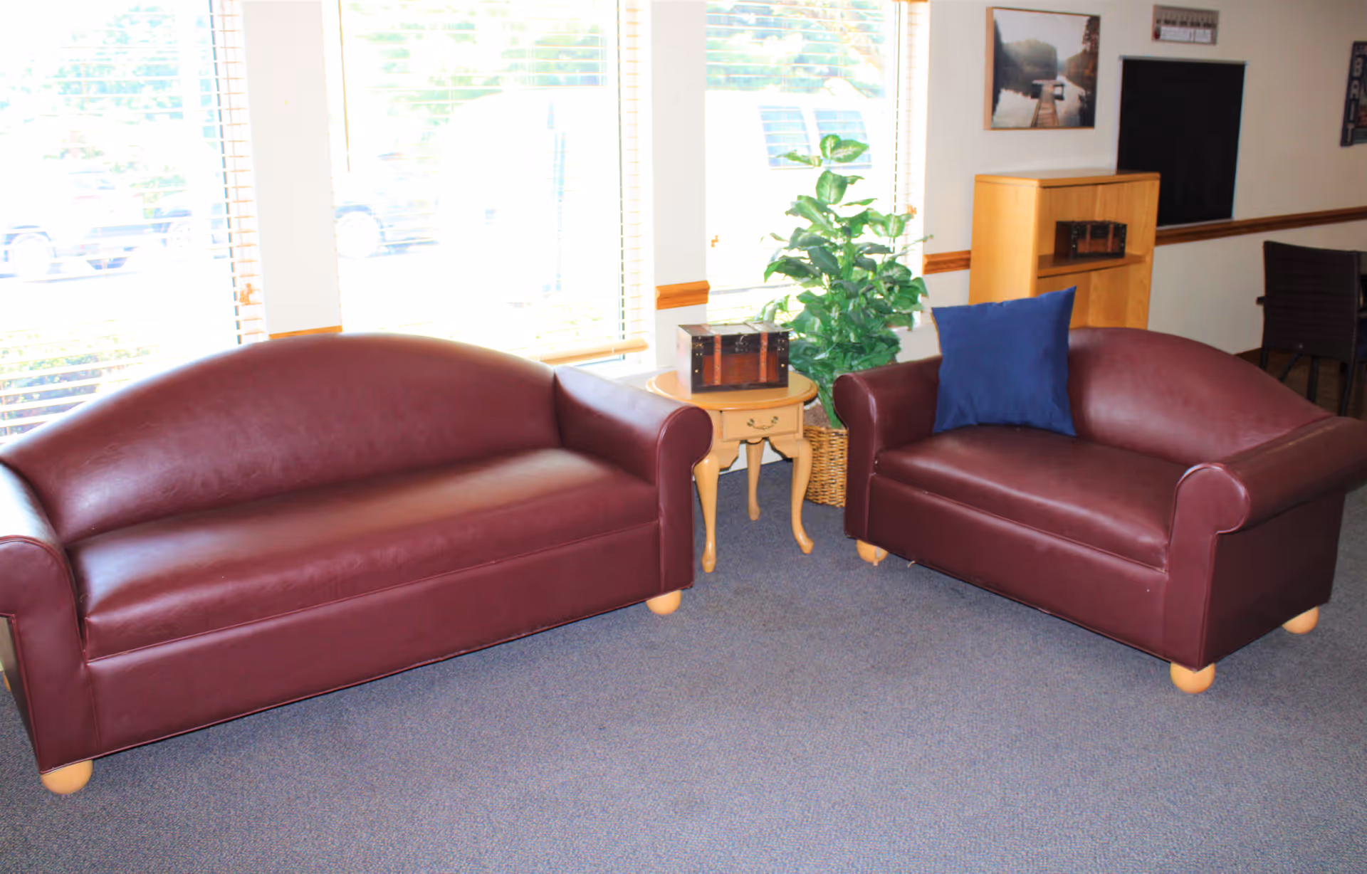 Two burgundy leather sofas facing each other in a sunlit lounge area with a small wooden side table and a potted plant.