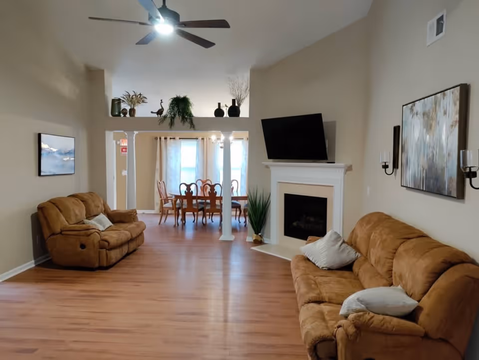 A spacious living room with two brown recliner sofas, a white fireplace with a mounted flat-screen TV above it, and a large painting on the wall. The room has wooden flooring, beige walls, and a ceiling fan with a light. In the background, there is a dining area with a wooden dining table and chairs, separated by white columns and decorated with plants and vases on a ledge above.