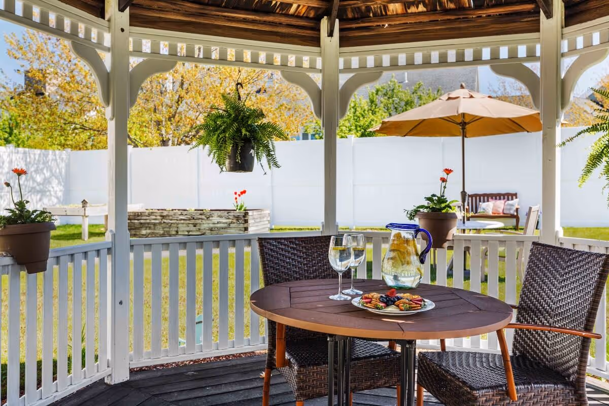 View from inside a white wooden gazebo overlooking a grassy backyard with a raised garden bed and a white fence. Inside the gazebo, there is a round wooden table with two wicker chairs. On the table, there is a pitcher of water with lemon slices, two glasses of water, and a plate of assorted crackers and berries. Hanging plants and potted flowers decorate the gazebo, and an outdoor umbrella shades a bench and table in the background.