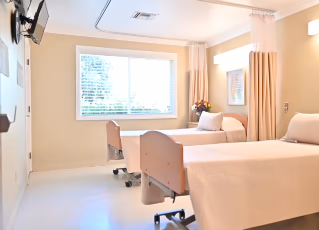 A bright healthcare room with two hospital beds covered in beige linens, a large window with blinds letting in natural light, a wall-mounted TV, and beige curtains separating the beds. There is a small bouquet of flowers on a table near the window.