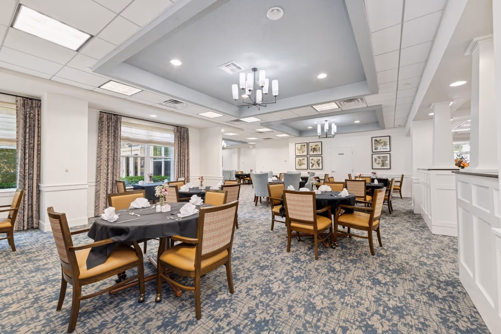 A spacious dining room in a senior living facility with multiple round tables covered with black tablecloths, each set with white folded napkins, silverware, and small floral centerpieces. The room features patterned carpet, large windows with curtains, white walls, and modern chandeliers hanging from a recessed ceiling.