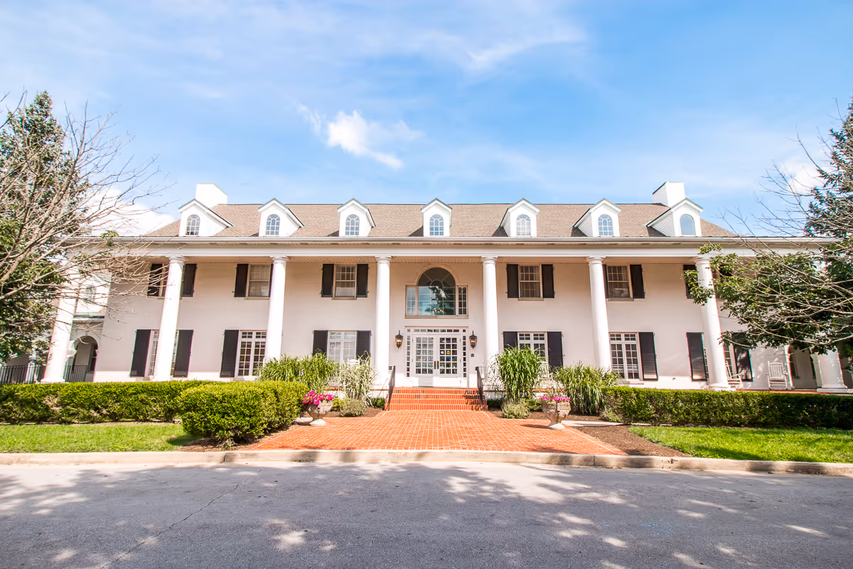 Front exterior view of a large white building with multiple windows, black shutters, and tall white columns. There is a brick walkway leading to the main entrance, flanked by green bushes and potted plants with flowers. The sky is blue with some clouds.