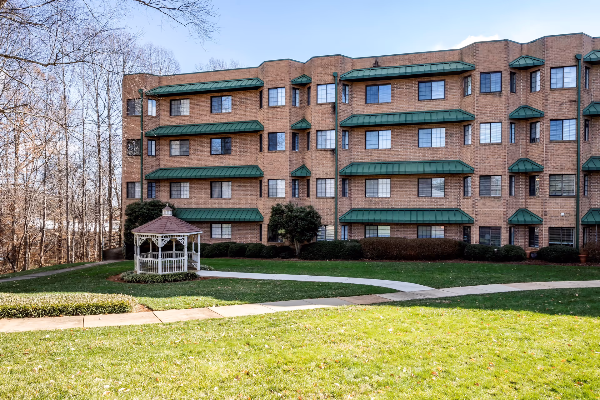 Exterior view of a multi-story brick building with green awnings over the windows. In front of the building is a well-maintained lawn with a white gazebo and a curved concrete walkway. Leafless trees are visible in the background under a clear sky.