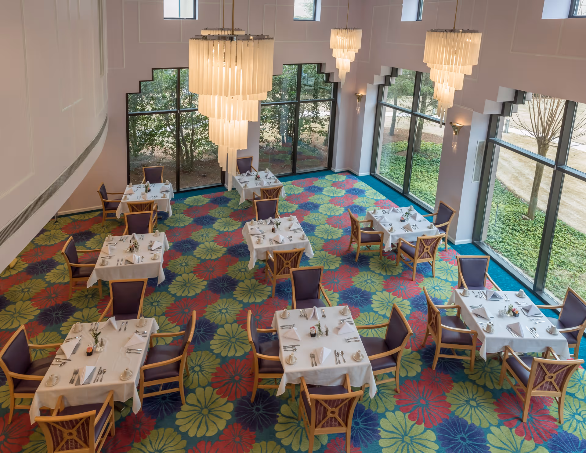 A bright dining room with large floor-to-ceiling windows letting in natural light. The room features colorful floral-patterned carpet, several tables covered with white tablecloths, each set with napkins, plates, cups, and silverware. Wooden chairs with purple cushions surround the tables. Three large, elegant chandeliers hang from the ceiling.