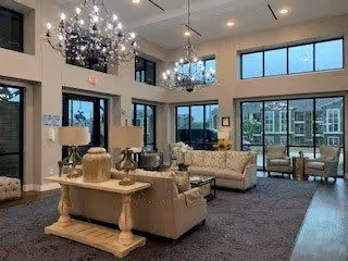 A spacious and well-lit living room area with large windows allowing natural light to fill the space. The room features comfortable beige sofas, a wooden coffee table with decorative vases, and two elegant chandeliers hanging from the ceiling. Outside the windows, a view of an apartment or residential building is visible.