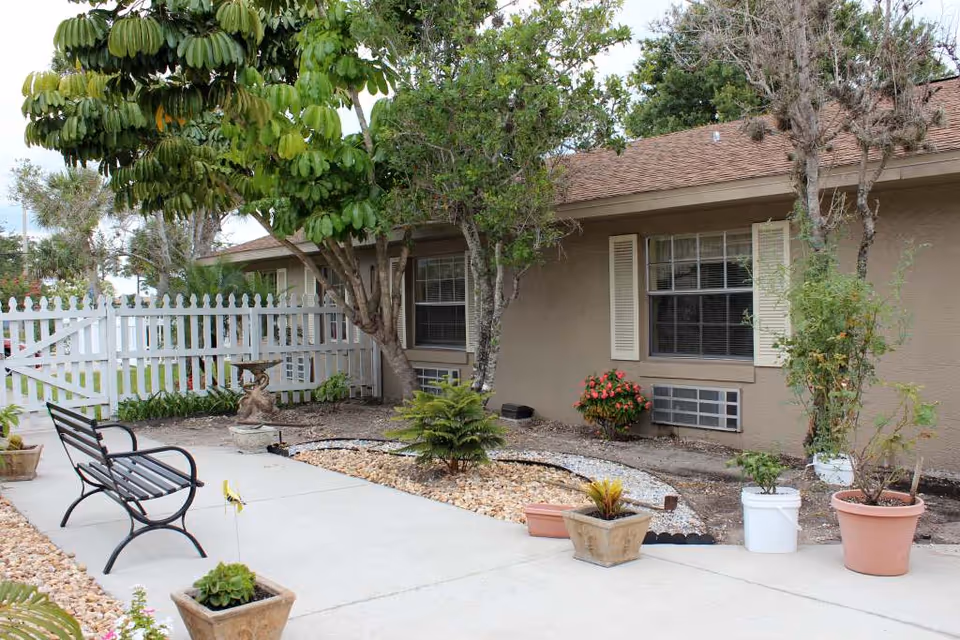 Outdoor garden area at Rockledge Health & Rehabilitation Center featuring a black metal bench on a concrete pathway, various potted plants, small trees, and a white picket fence in the background next to a beige building with windows and shutters.