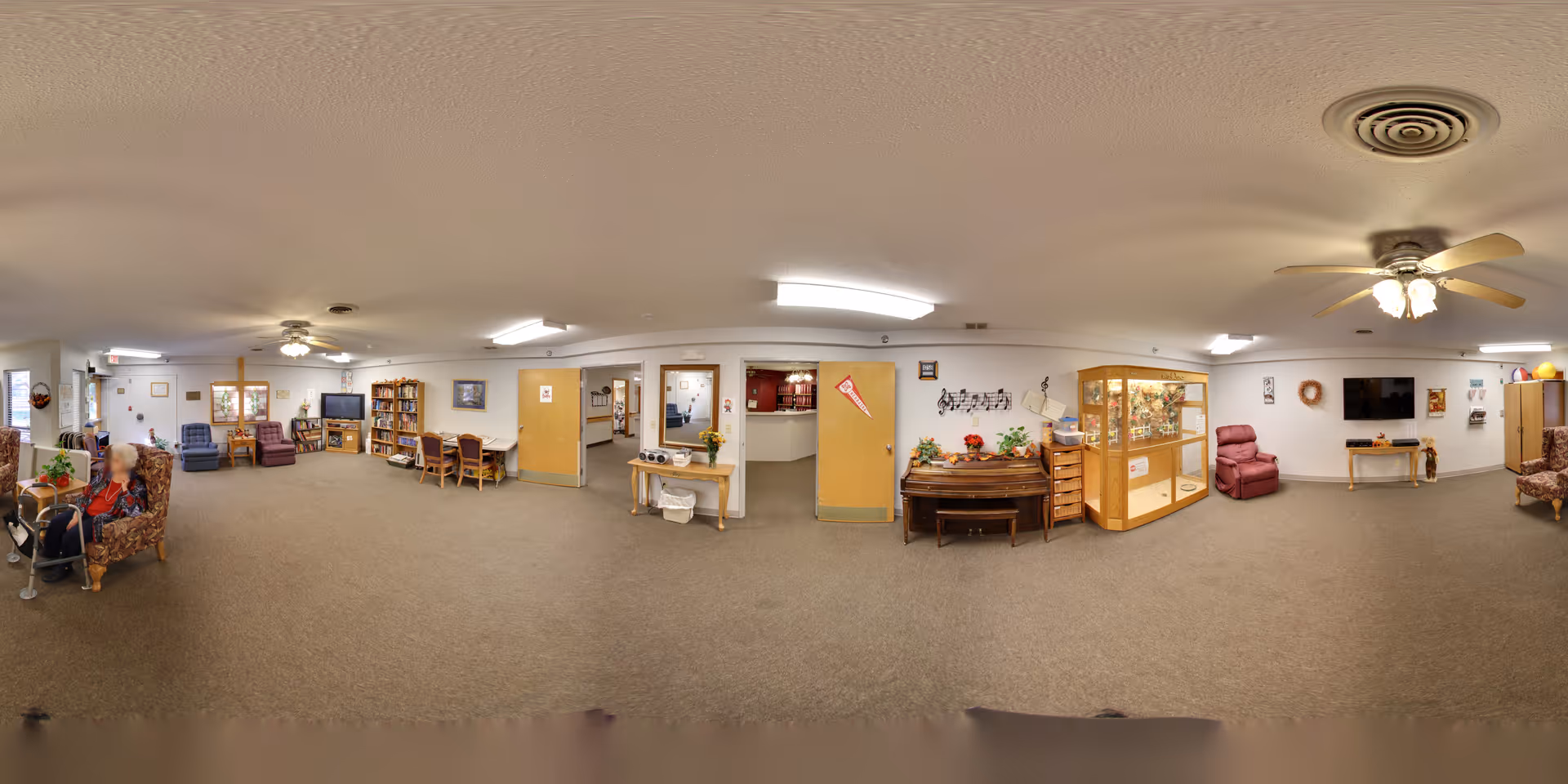 Carpeted communal lounge with armchairs, recliners, bookshelves, a TV, tables, and display cabinets.