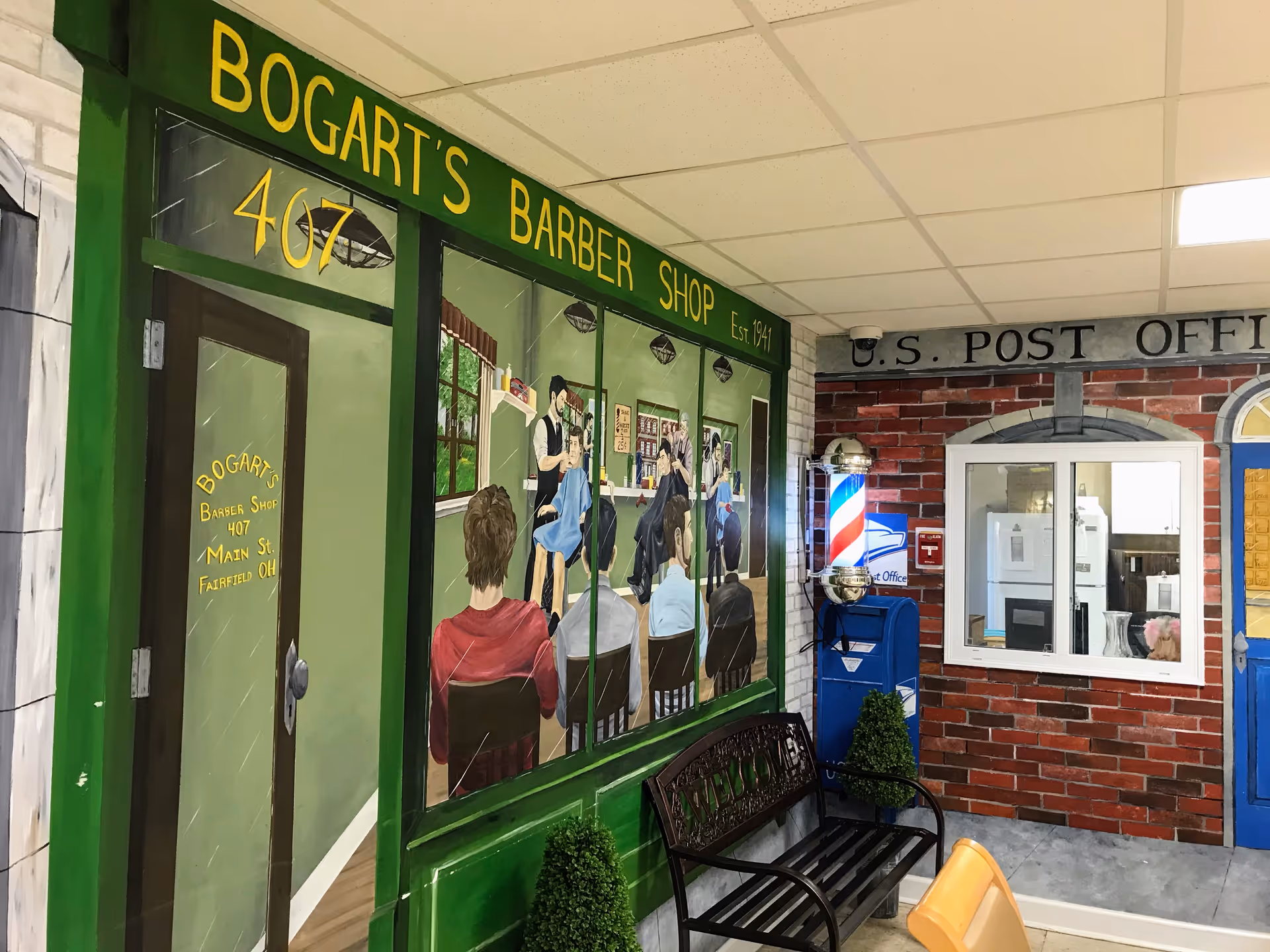 Interior view of a senior living facility area decorated to resemble a small town street scene, featuring a mural of Bogart's Barber Shop with people getting haircuts, a bench in front, a U.S. Post Office facade with a mailbox, and a barber pole light fixture.