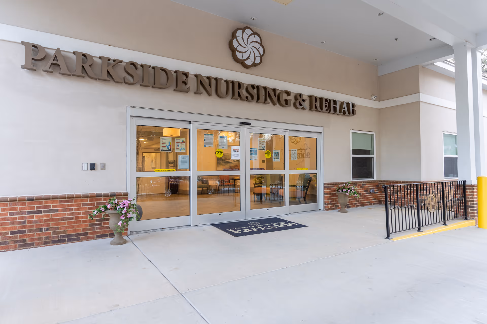Entrance to Parkside Nursing & Rehab facility with automatic sliding glass doors, two flower pots on either side, a black railing ramp, and a mat with the Parkside logo on the ground.