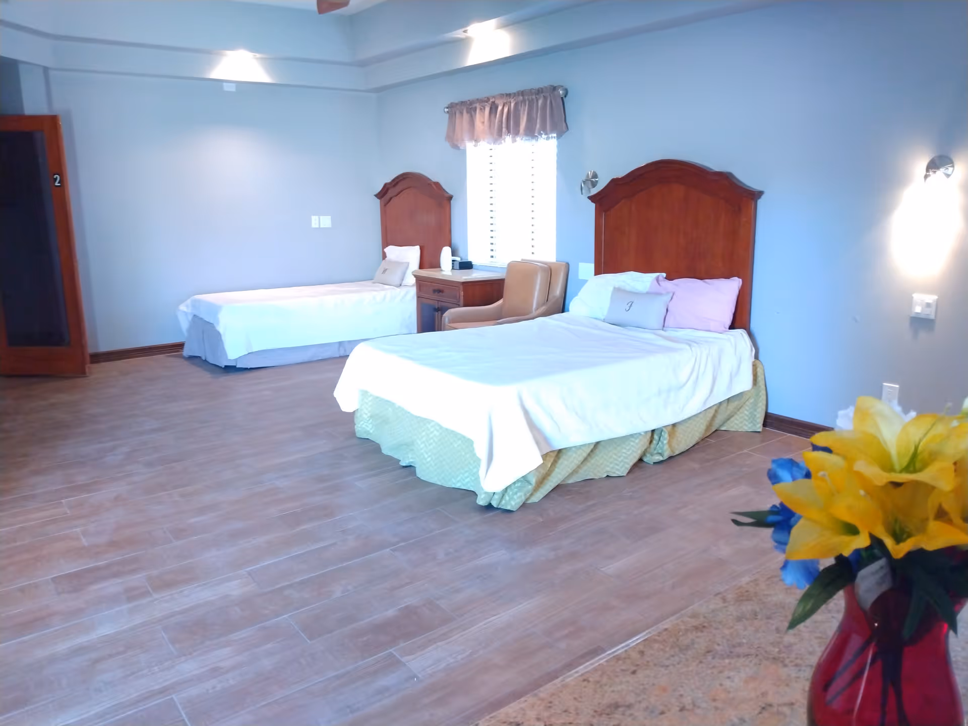 A spacious bedroom with two single beds, wooden headboards, a nightstand and chair, and a vase of yellow flowers in the foreground.