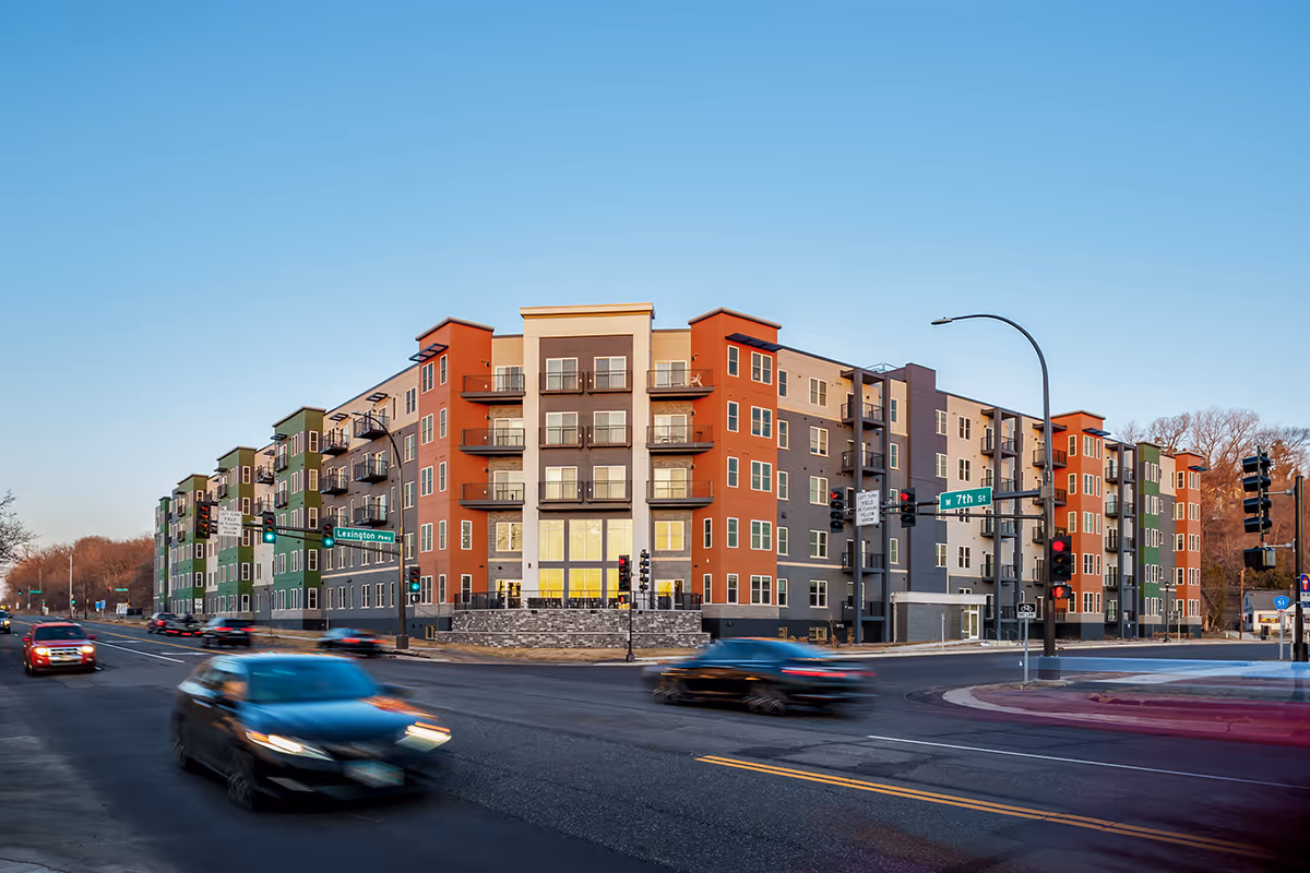 A modern multi-story residential building with colorful exterior walls in shades of green, red, gray, and beige, located at the intersection of Lexington Parkway and W 7th Street. Several cars are driving on the street in front of the building under a clear blue sky.
