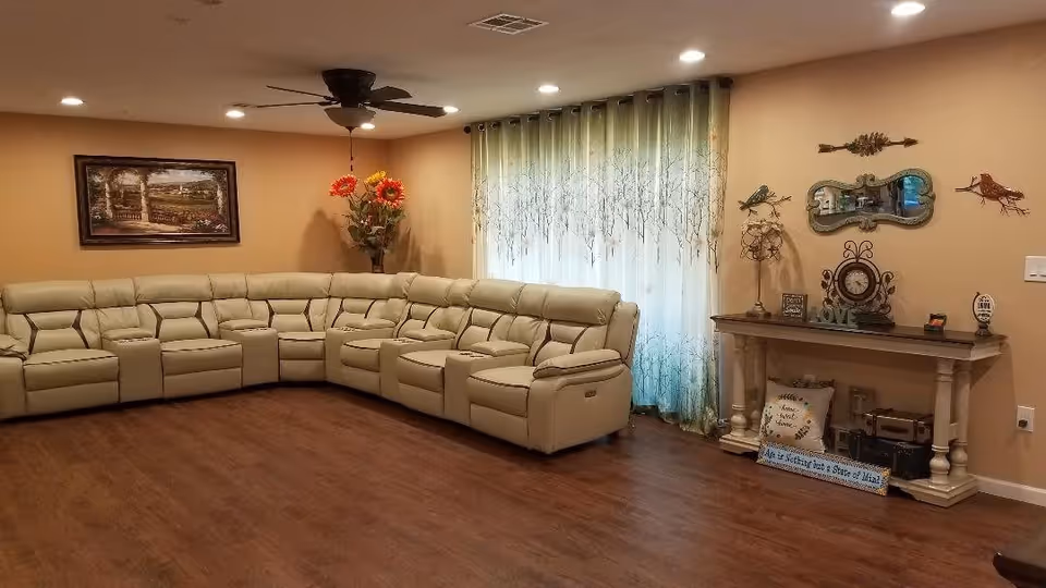A spacious living room with a large beige sectional recliner sofa arranged in an L-shape. The room features wooden flooring, beige walls, a ceiling fan with lights, and a window covered with sheer curtains with a tree branch pattern. On one wall, there is a framed landscape painting, and on another wall, a decorative console table with various ornaments including a clock, a small mirror, and bird wall decorations.