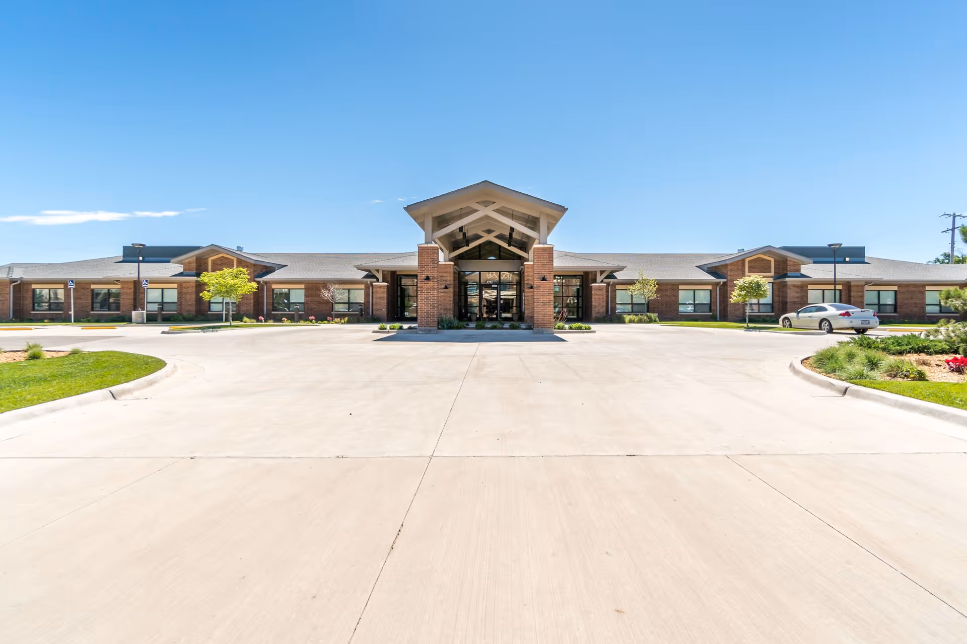 Front exterior view of a single-story brick building with a covered entrance, surrounded by a paved driveway and landscaped greenery under a clear blue sky.