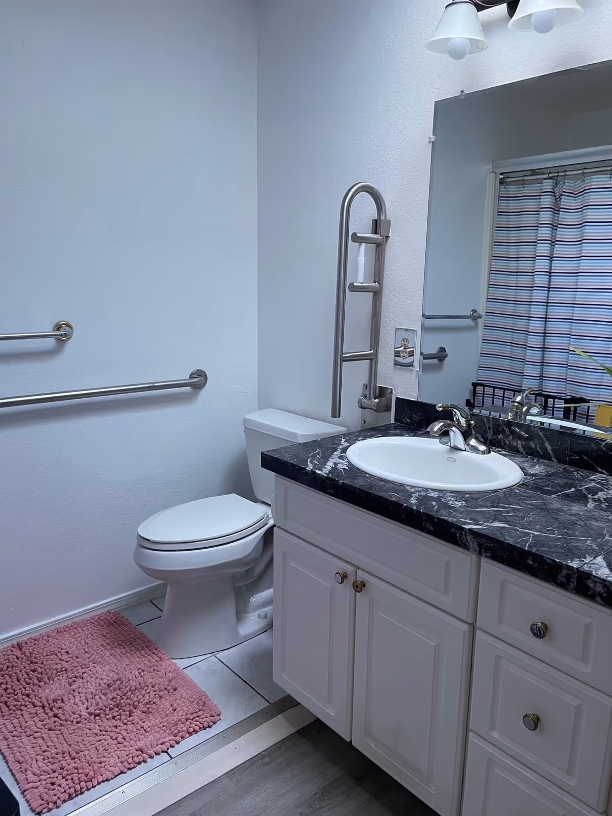 A bathroom with a white toilet, white vanity with a black marble countertop, a white sink, and a large mirror above the sink. There are two metal grab bars on the wall near the toilet, a pink textured bath mat on the tiled floor, and a striped shower curtain visible in the mirror reflection.