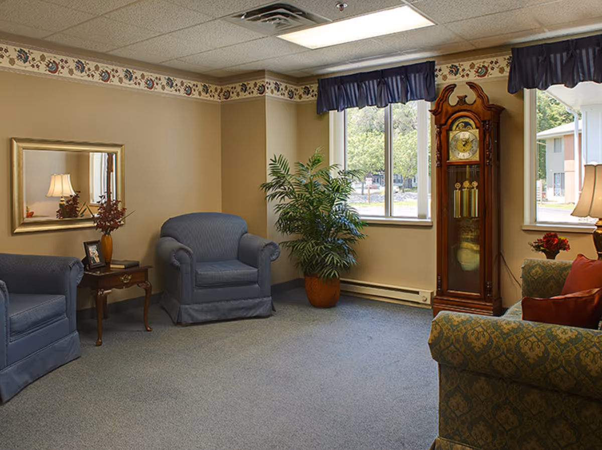 A cozy living room with two blue upholstered armchairs, a wooden side table with a lamp and decorative items, a large potted plant near two windows with blue valances, and a tall wooden grandfather clock. The walls are beige with a floral border near the ceiling.