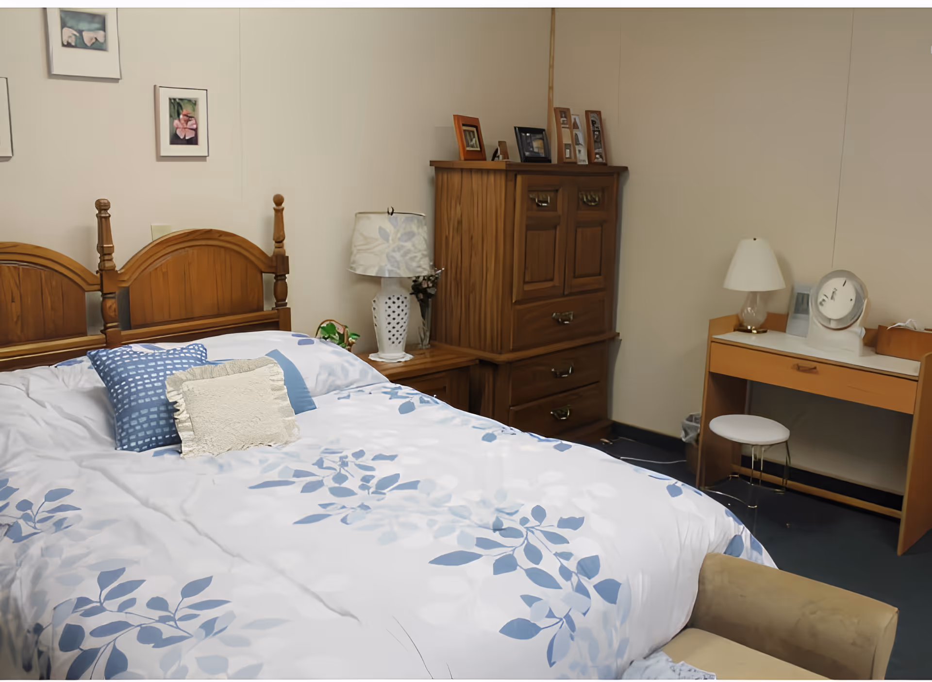 A neatly made bedroom with a wooden headboard bed covered in blue floral bedding, a wooden dresser and nightstand with lamps, and a small desk.
