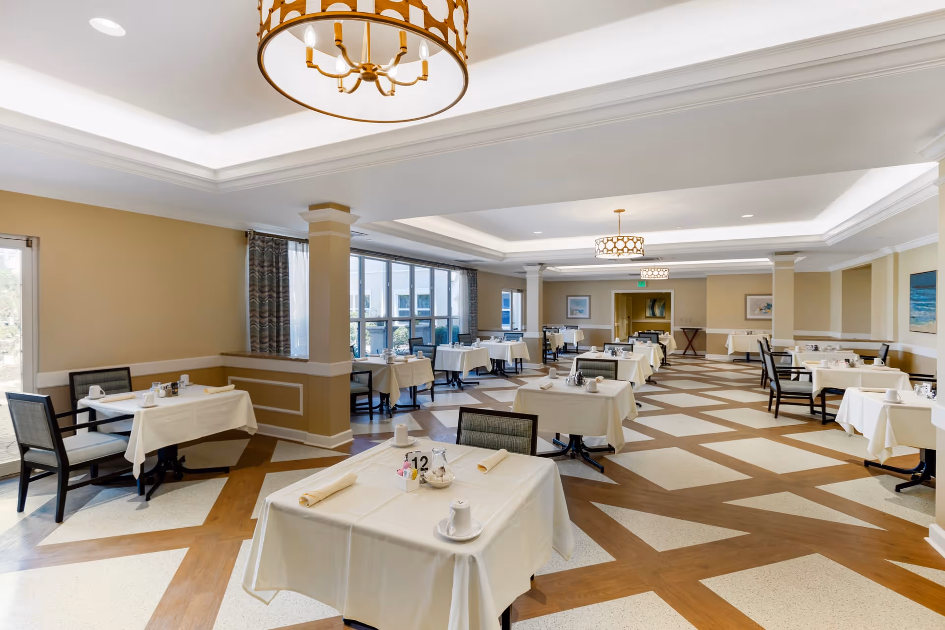 A spacious dining room with multiple tables covered in white tablecloths, each set with cups, napkins, and condiments. The room features large windows with curtains, beige walls, decorative ceiling lights, and a patterned floor with wood and light-colored tiles.