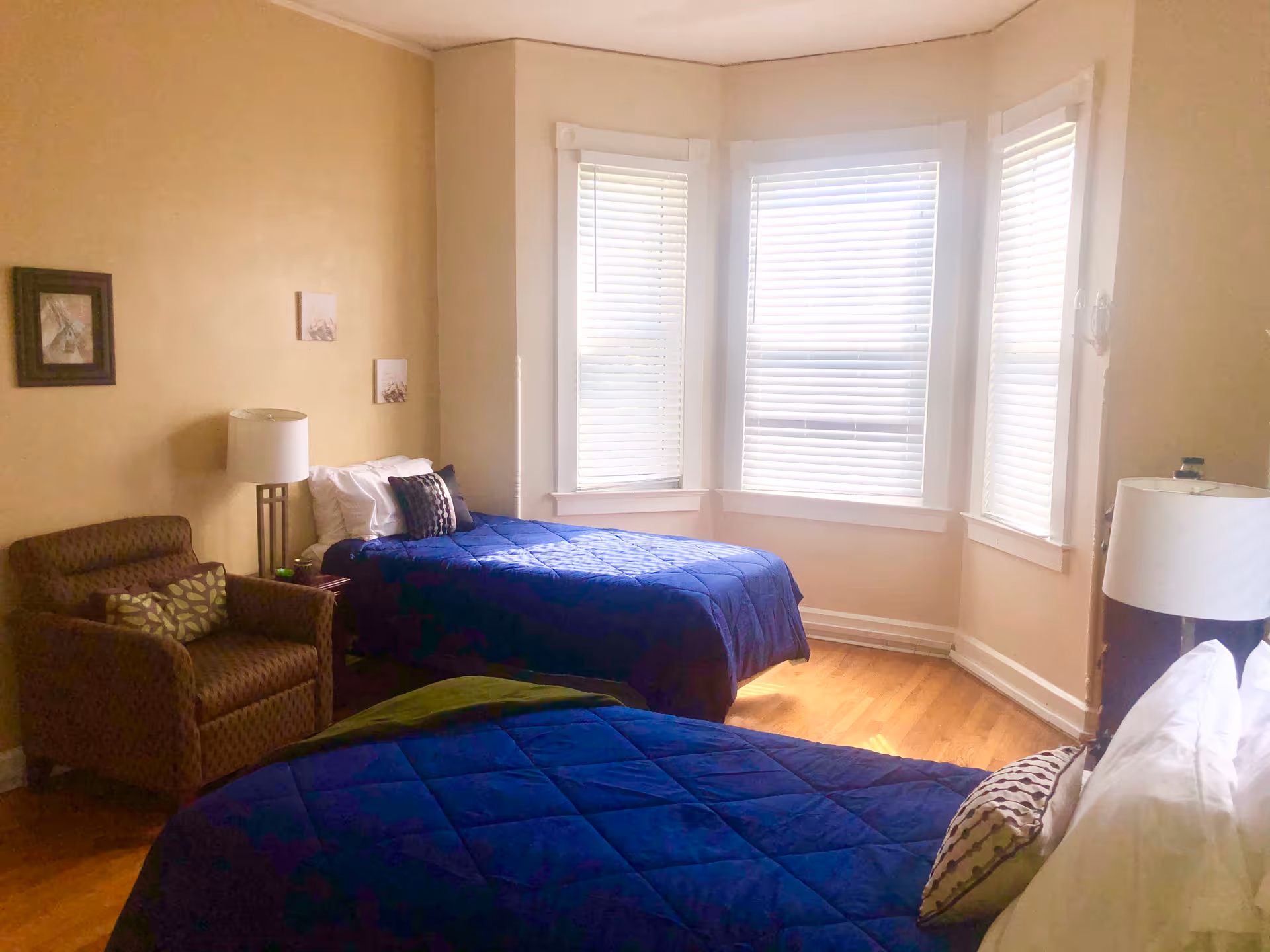 A bedroom with two twin beds covered in blue comforters, each with pillows. There is a brown armchair with a patterned cushion next to a small table with a lamp. The room has three large windows with white blinds and light-colored walls.