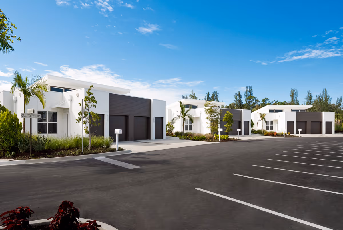 Modern single-story white buildings with dark garage doors, surrounded by small trees and plants, under a blue sky with scattered clouds. There is a parking lot with marked spaces in the foreground and a street sign labeled 'Calistoble Loop'.