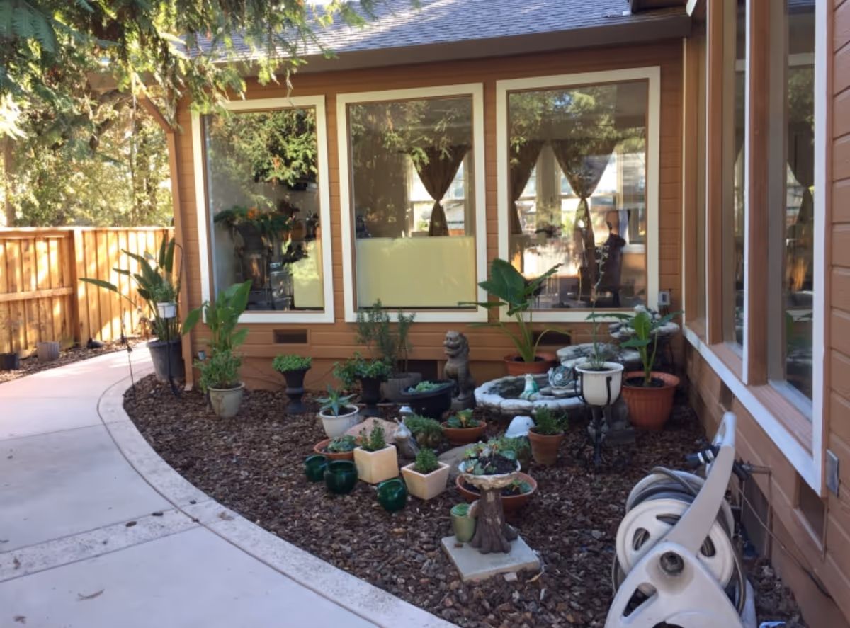 Outdoor garden area next to a building with large windows. The garden features various potted plants and small decorative statues arranged on mulch. A curved concrete pathway runs alongside the garden, and a wooden fence is visible in the background.