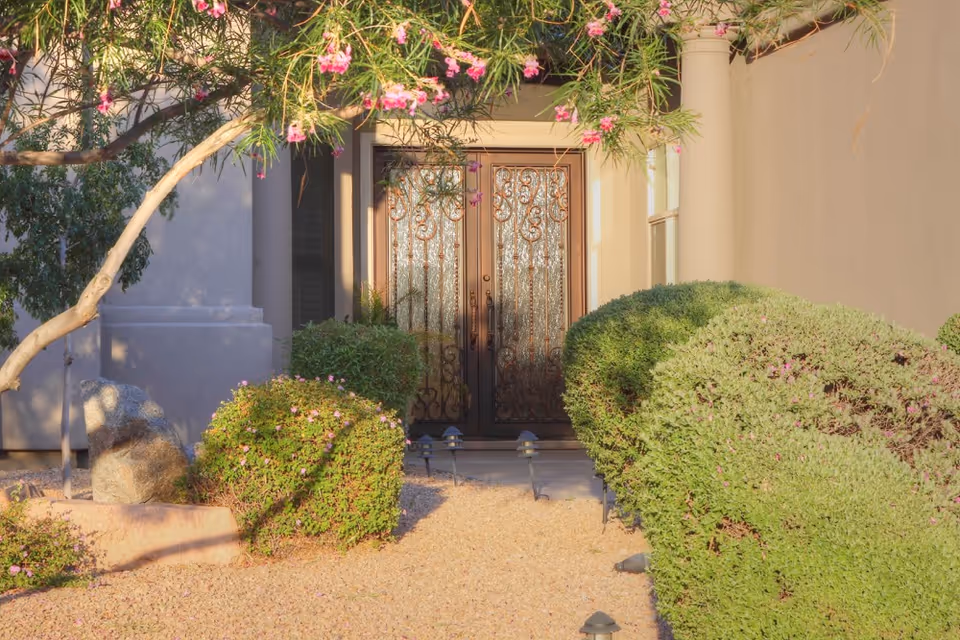 Front entrance of a building with decorative double doors, surrounded by neatly trimmed bushes and a tree with pink flowers. The pathway leading to the door is covered with small gravel.