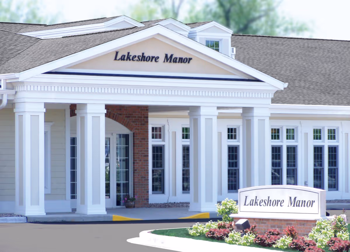 Front exterior view of Lakeshore Manor building with white columns, large windows, and a sign in front surrounded by landscaping.