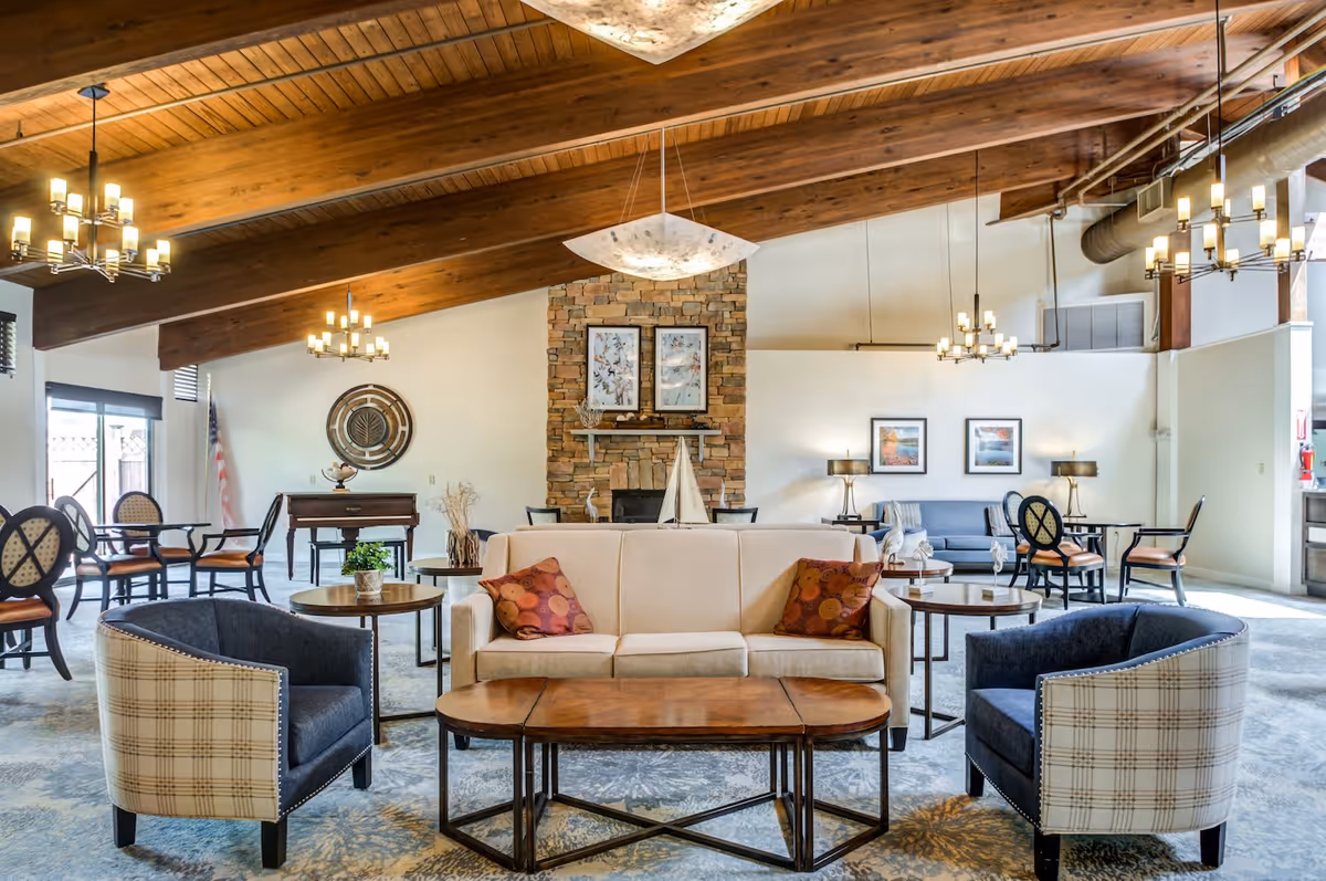 A spacious and well-lit common area with a high wooden ceiling and exposed beams. The room features a beige sofa with two patterned pillows, two plaid and navy armchairs, and several wooden tables. In the background, there is a stone fireplace with framed artwork above it, additional seating areas with chairs and tables, and decorative lighting fixtures hanging from the ceiling.
