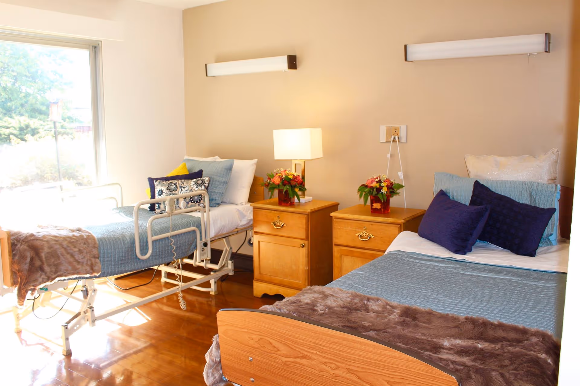 A bright and clean room with two beds, one hospital-style bed with safety rails and one regular bed, both made with blue bedding and decorative pillows. Between the beds are two wooden nightstands with flower arrangements and a lamp. A large window lets in natural light, illuminating the polished wooden floor.