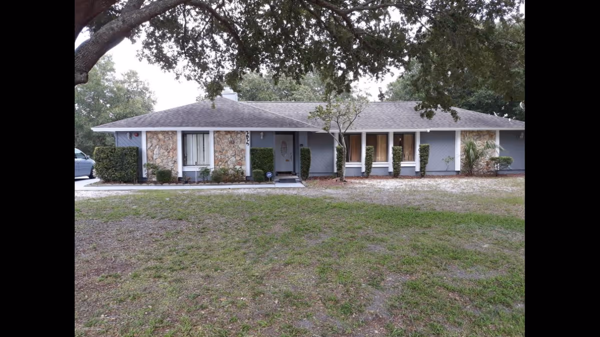 Single-story residential building with a gray roof and stone accents on the walls, surrounded by trimmed bushes and trees, with a grassy front yard and a car partially visible on the left side.