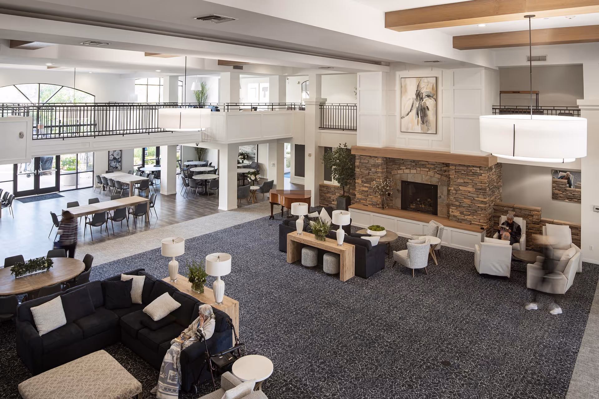 Spacious senior living facility common area with a large stone fireplace, multiple seating arrangements including sofas and armchairs, a piano, and dining tables in the background. The room has high ceilings, large windows, and modern lighting fixtures. Two elderly individuals are seated in the lounge area.