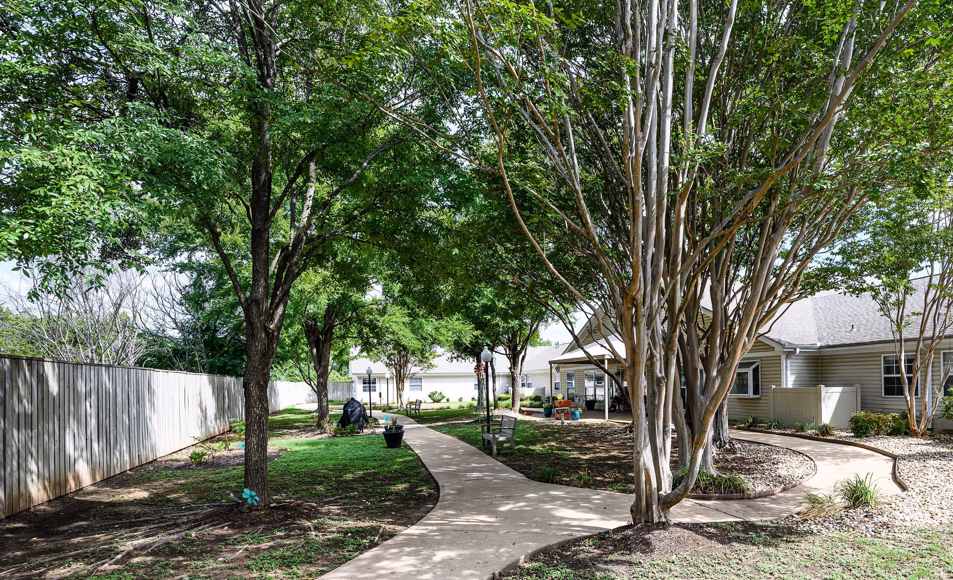 A peaceful outdoor garden area at Arden Courts - ProMedica Memory Care Community in Austin featuring a paved walkway winding through green grass and trees. There are benches along the path and residential buildings visible in the background under a partly cloudy sky.