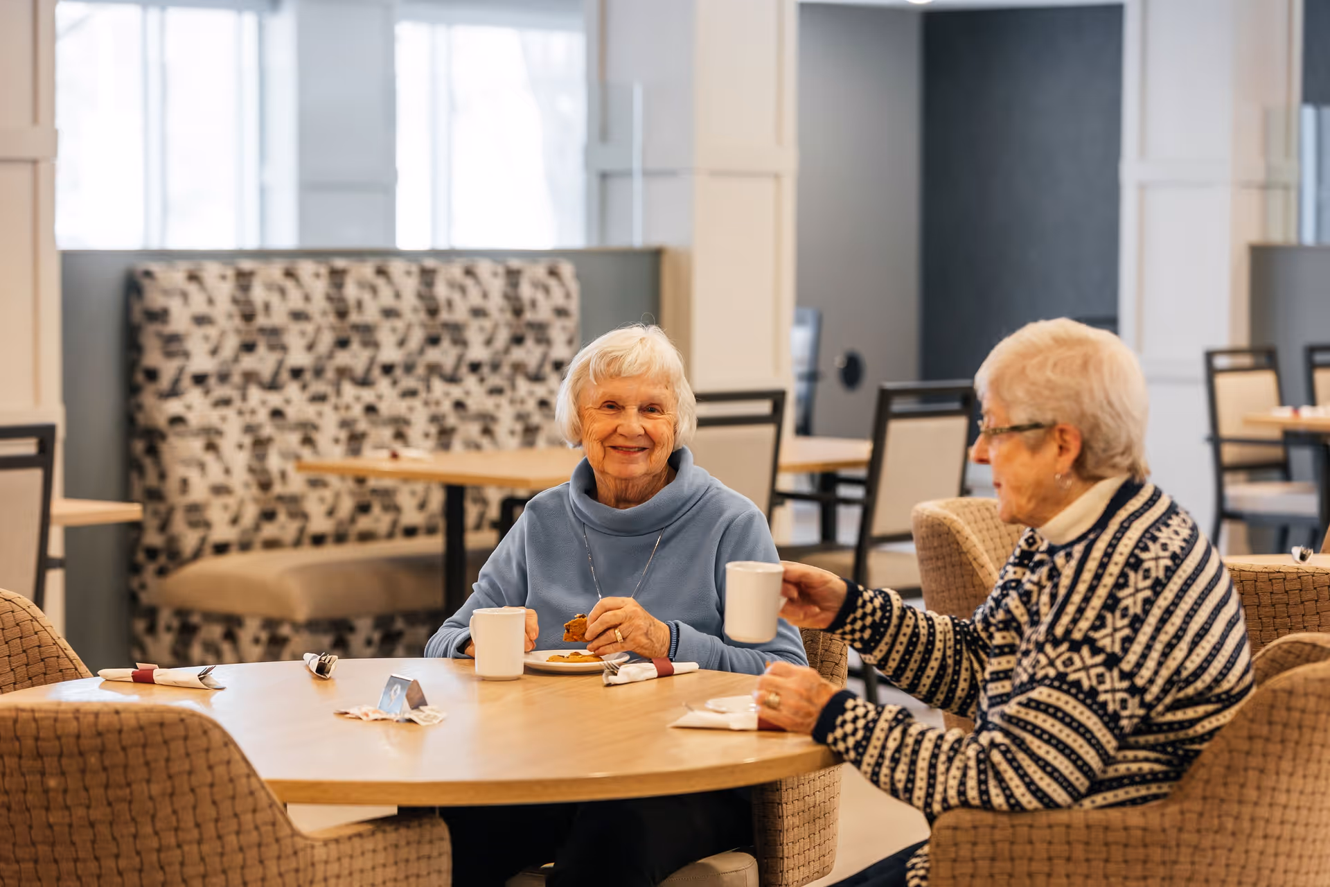 Two elderly women sitting at a round wooden table in a dining area, enjoying coffee and cookies. One woman is smiling at the camera while holding a cookie, and the other is raising a white mug. The background shows upholstered seating and additional tables and chairs in a well-lit room.