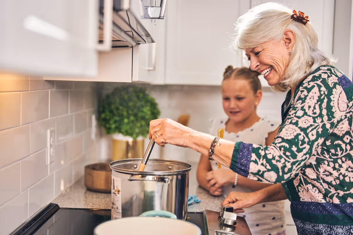 An elderly woman with white hair and a floral patterned shirt is stirring a pot on a stove in a kitchen while smiling. A young girl stands beside her, watching and smiling. The kitchen has white cabinets and a light-colored tiled backsplash.