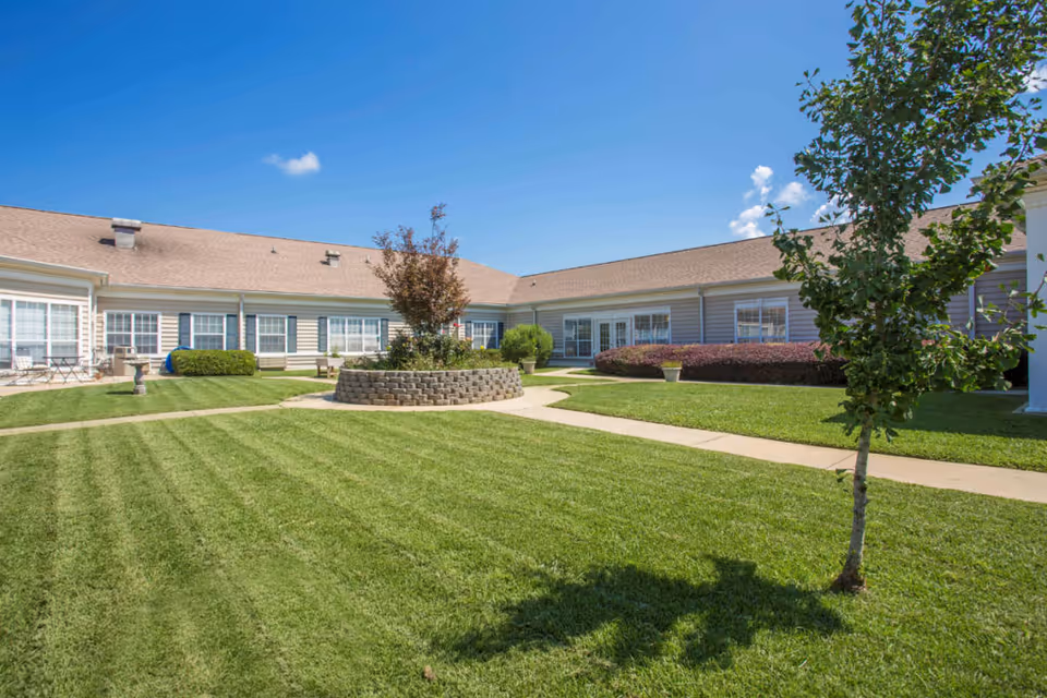 Sunlit courtyard with a manicured lawn, paved walkways, a central raised planter, and single-story building wings surrounding it.