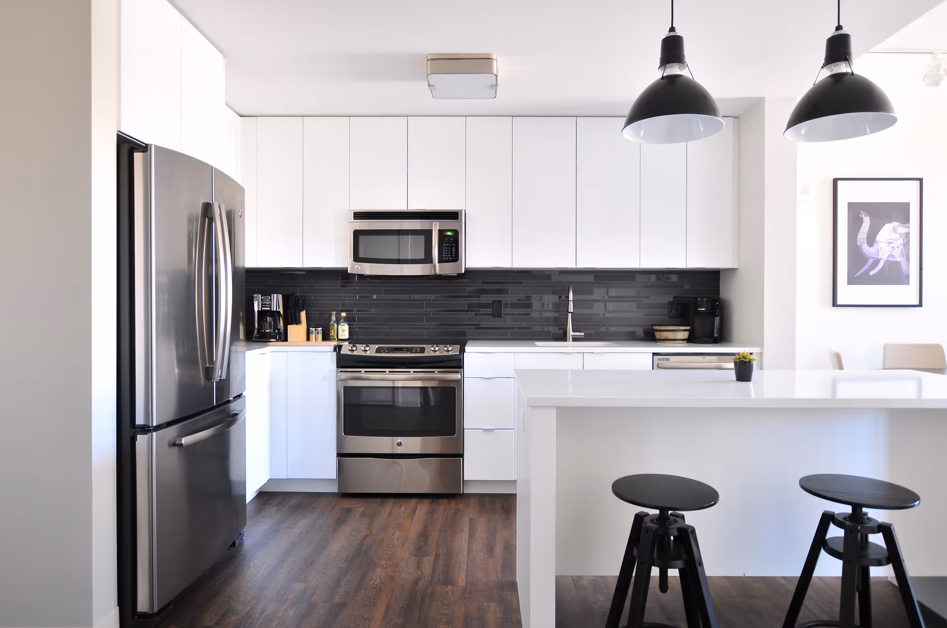 Modern kitchen with white cabinets, stainless steel refrigerator, oven, microwave, and dishwasher. The kitchen features a black tile backsplash, a white island with two black stools, two black pendant lights hanging from the ceiling, and a framed artwork on the wall.