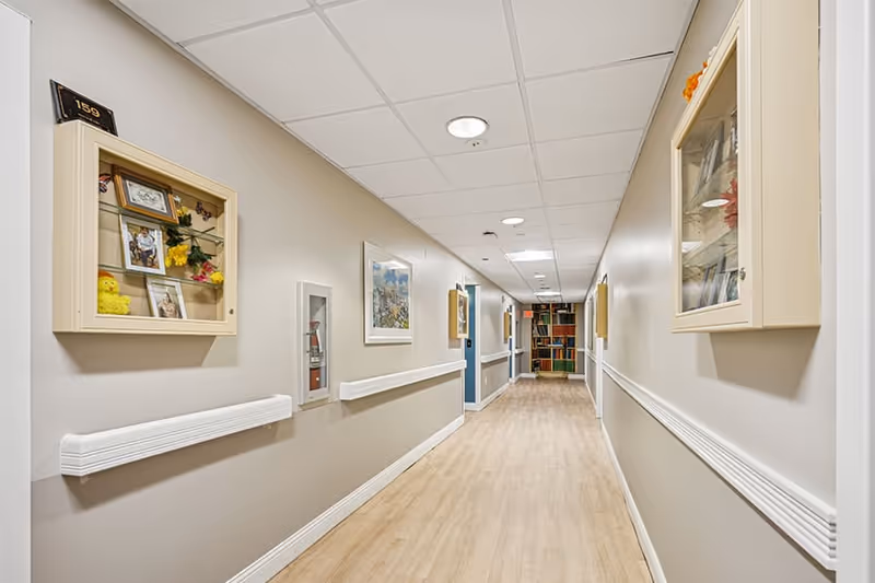 A well-lit hallway in a senior living facility with light wood flooring and beige walls. The walls have white handrails and display cases containing photos and decorative items. At the end of the hallway, there is a colorful bookshelf filled with books.