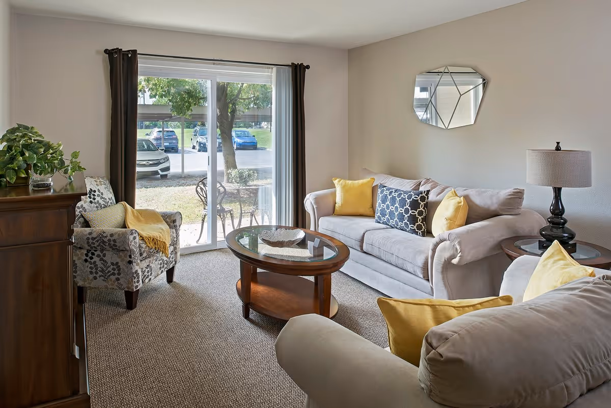 A cozy living room in a senior living facility featuring a beige sofa with yellow and patterned cushions, a matching armchair with a yellow throw, a wooden coffee table with a glass top, a side table with a lamp, a decorative mirror on the wall, and a sliding glass door leading outside with a view of parked cars and greenery.