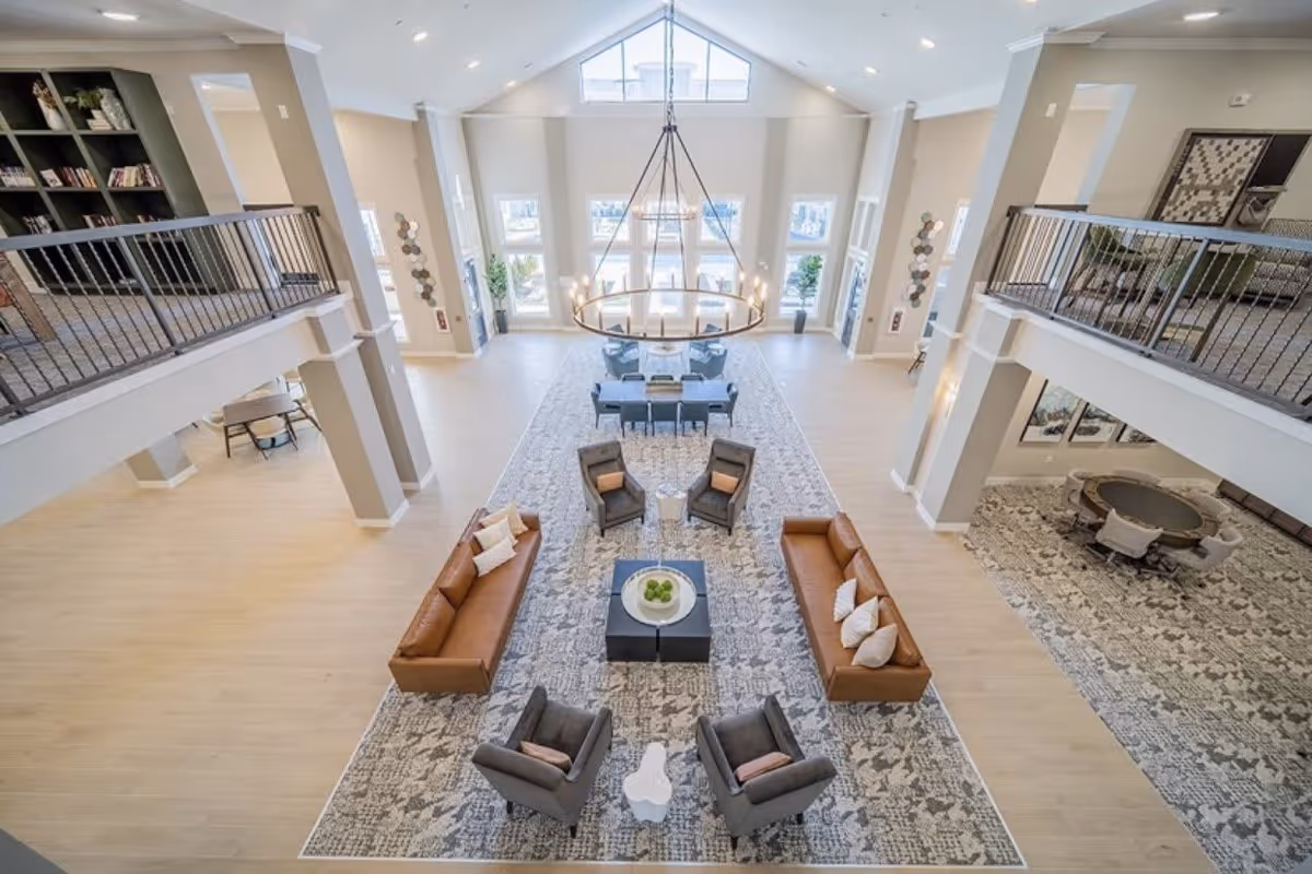 Spacious and bright senior living facility common area with high ceilings and large windows. The room features two brown leather sofas, four gray armchairs arranged around a central coffee table with a bowl of green apples, and a dining table with chairs near the windows. There is a large circular chandelier hanging from the ceiling and a patterned rug covering the floor. The area includes a second-floor balcony with railings and bookshelves visible on the upper level.