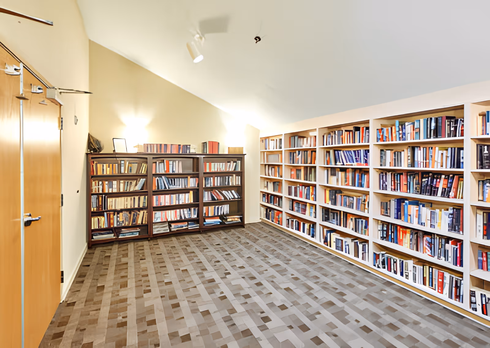 A quiet room with beige walls and a patterned carpet floor, featuring multiple bookshelves filled with books along the right wall and the back wall. Two wall-mounted lights illuminate the space, and there is a closed wooden door on the left side.